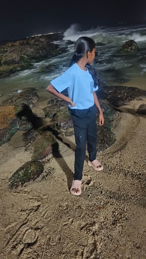Aradhya standing beside the rocky shoreline and powerful sea at Kanyakumari at night